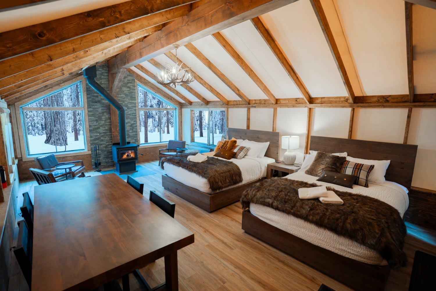 Lodge-style bedroom with two beds, wood stove, dining table and snowy forest view.