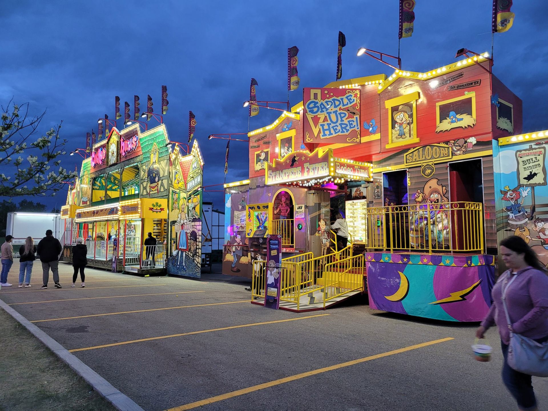 Two brightly lit carnival attractions, one with a "Saddle Up" cowboy theme, at dusk with people walking.