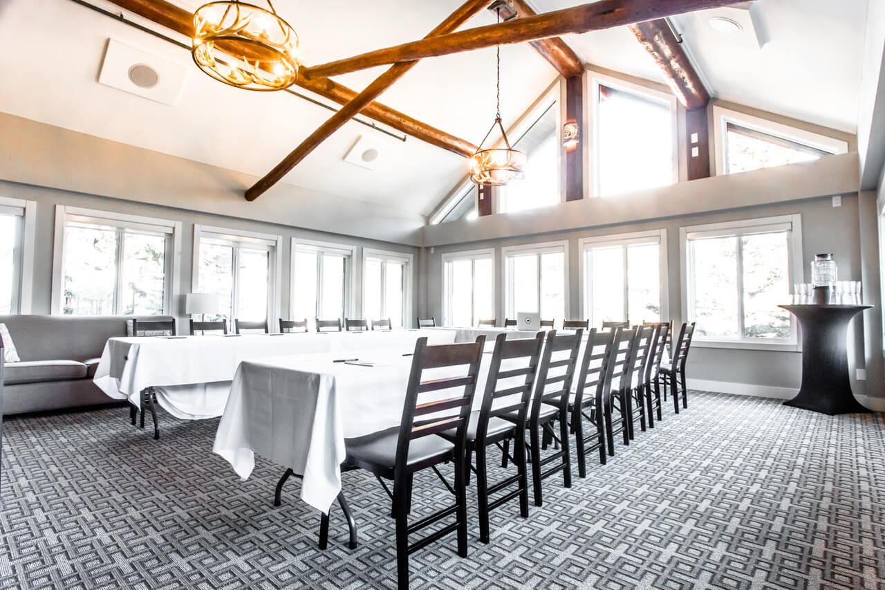 Large meeting room with U-shaped tables, wooden beams, bright windows, and modern lighting.