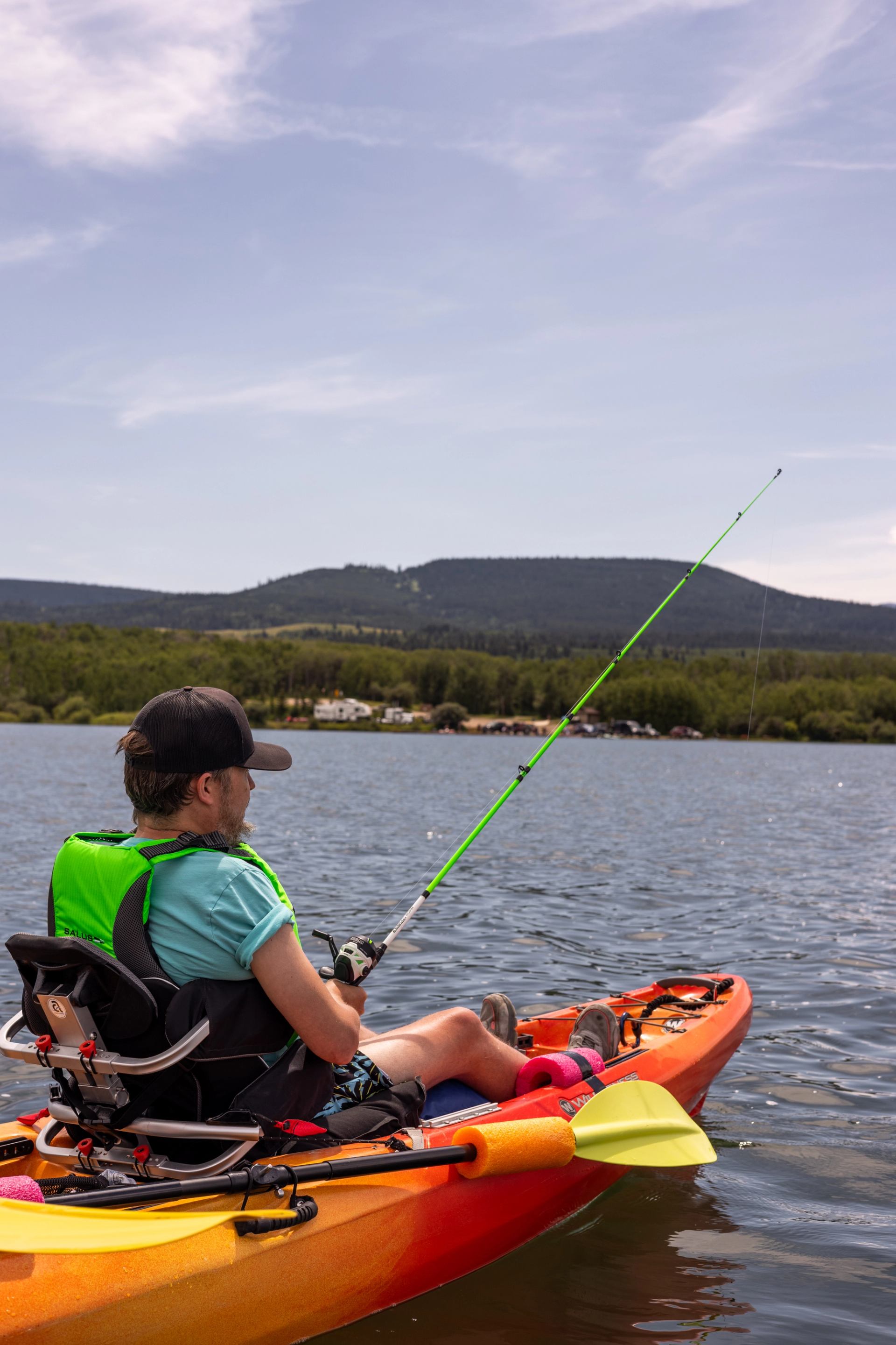 Person fishing from an adaptive kayak on a calm lake with hills in the background.