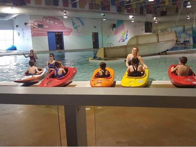 Group seated on colorful kayaks beside a large indoor swimming pool with slide.