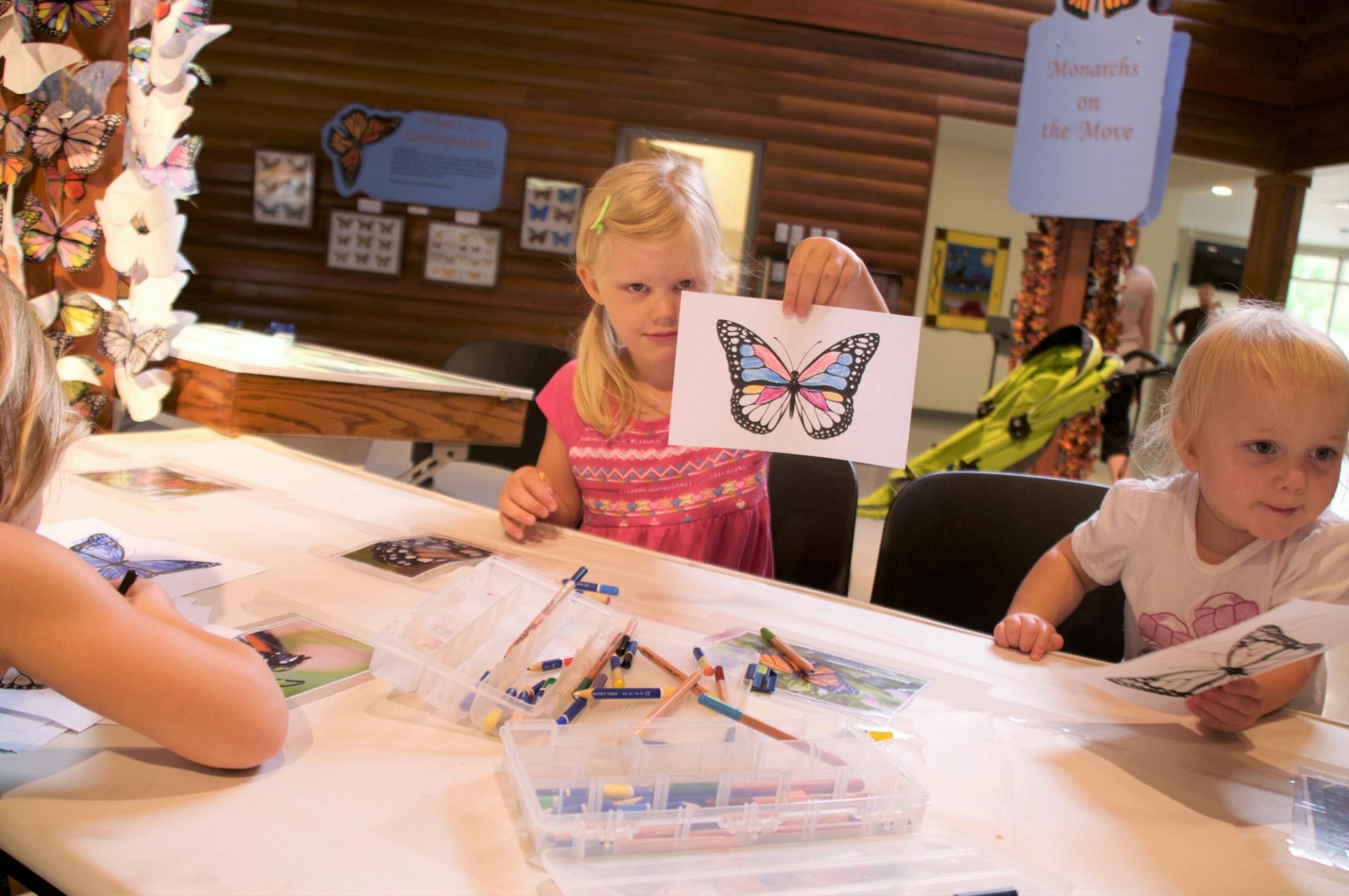 A child holds up a coloured butterfly drawing while sitting at a craft station.