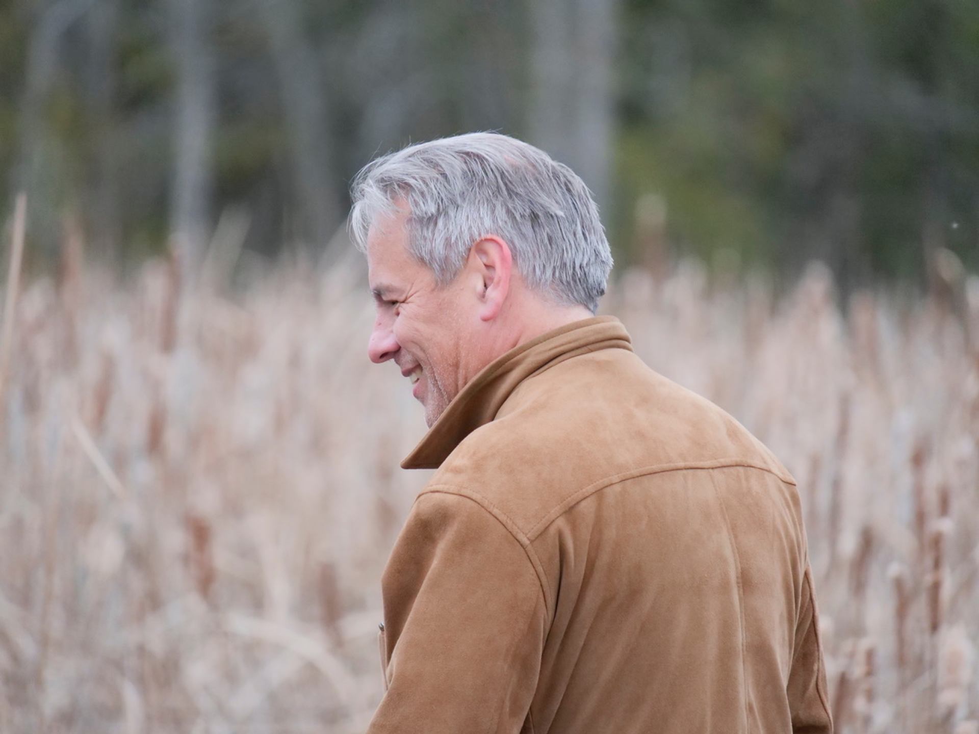 Smiling man in a brown jacket looking left, against blurred dry reeds.