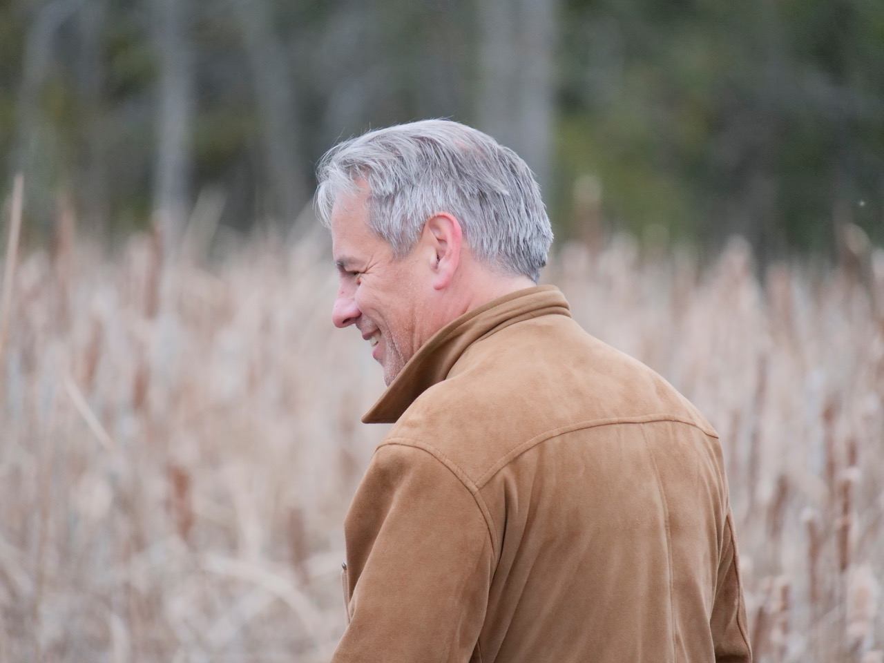 Smiling man in a brown jacket looking left, against blurred dry reeds.