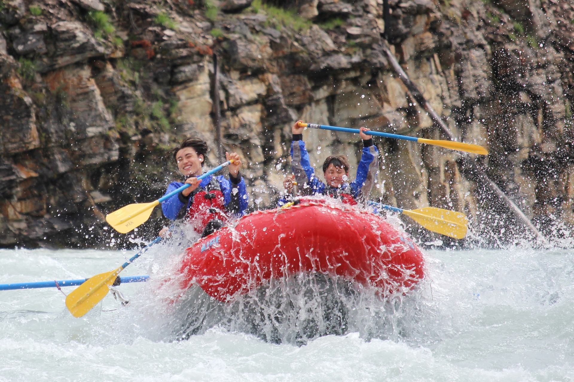 Smiling people in a red raft navigating splashing white water rapids.