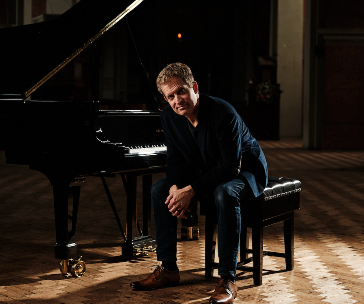 Person in dark blazer sits on piano bench beside grand piano in dim room.