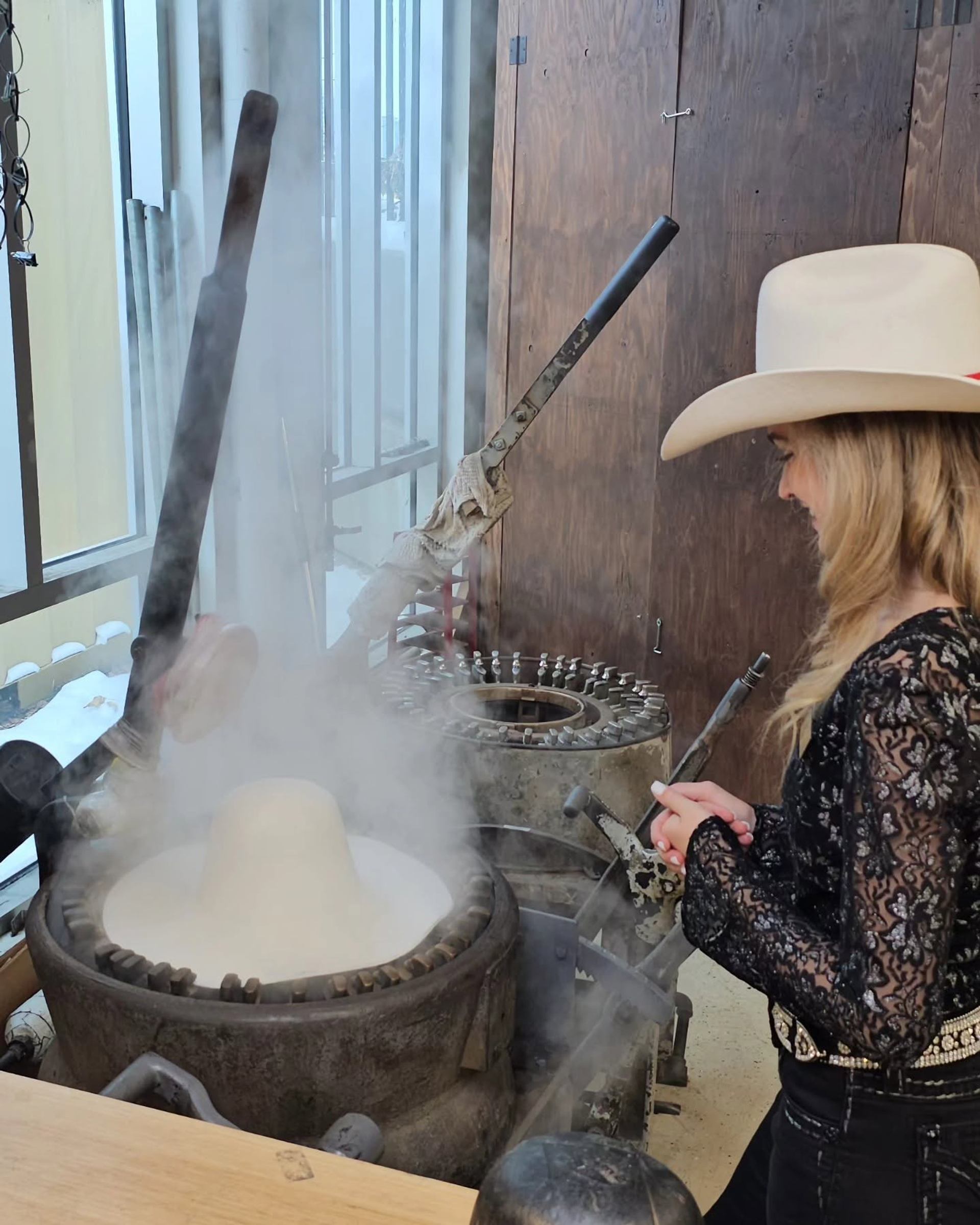 Person in cowboy hat watches steam rise from hat-shaped moulding machine indoors.