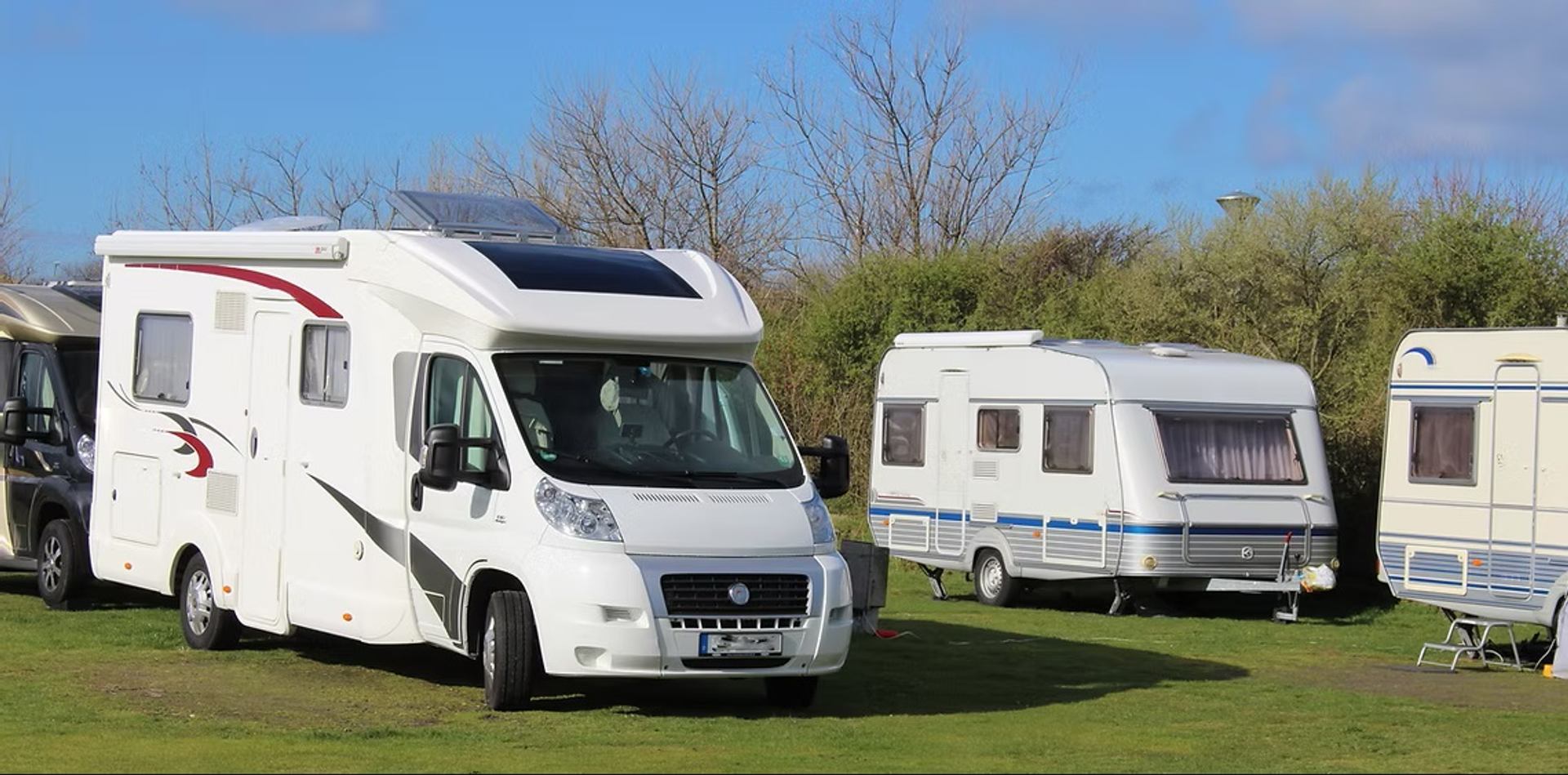 Several RVs are parked on grassy sites bordered by trees beneath a bright blue sky.