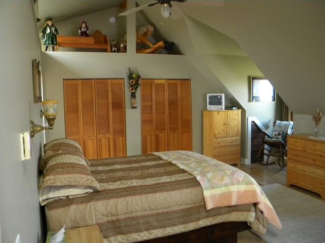 Bedroom interior with wooden wardrobe, bed, dresser, window, and ceiling fan in an attic room.