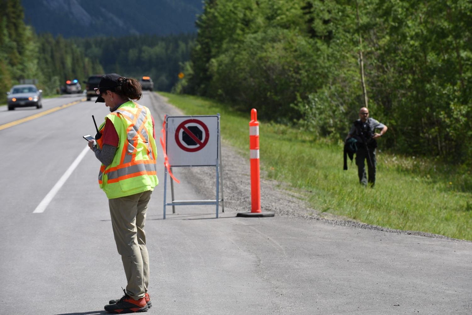 High-visibility runner walks along a road beside race volunteers and a vehicle convoy.