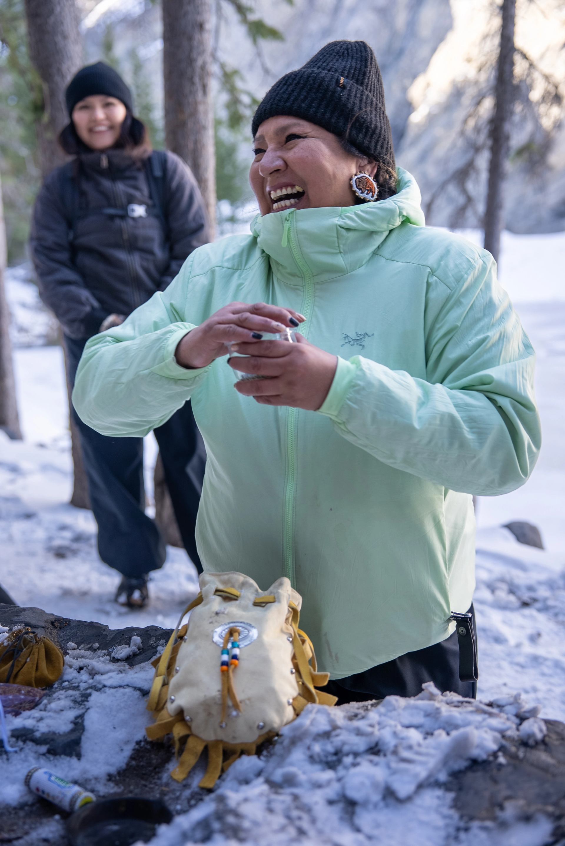 Person in mint jacket stands in snowy forest beside beaded leather pouch on rock.
