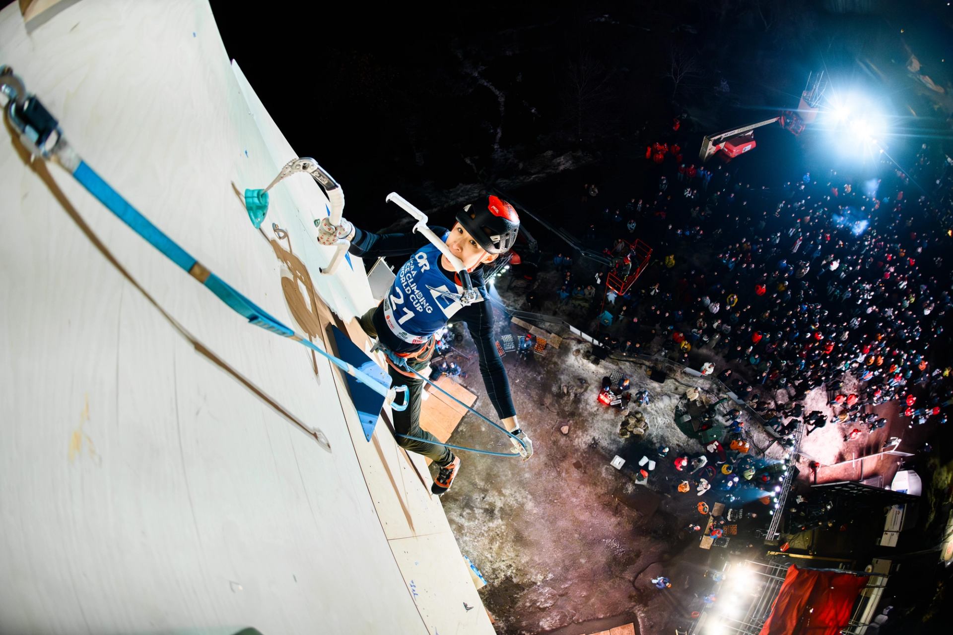 Ice climber on lit wall with crowd below at nighttime competition.