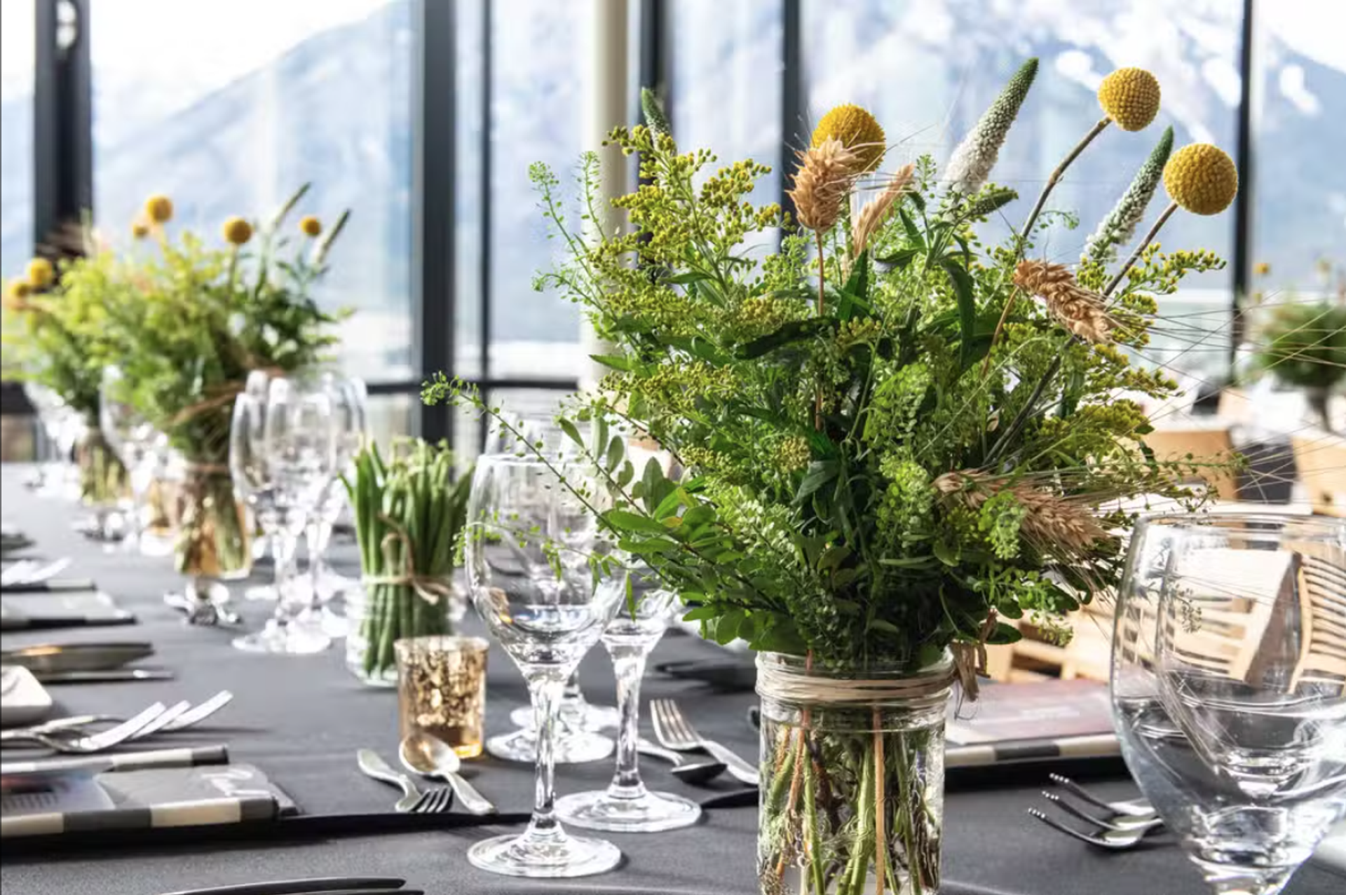 Table setting with green floral centrepieces in jars and wine glasses by large windows.