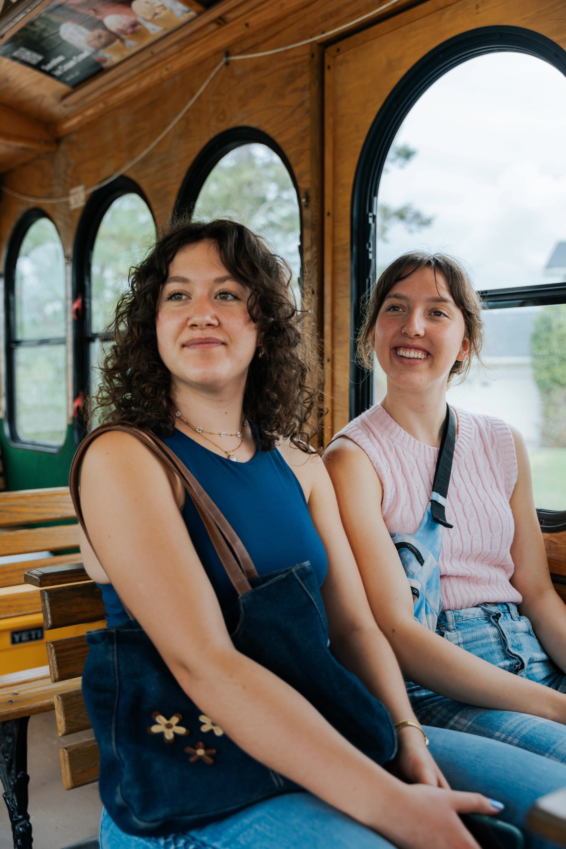 Two people sit on wooden trolley seats with bags, near large arched windows.