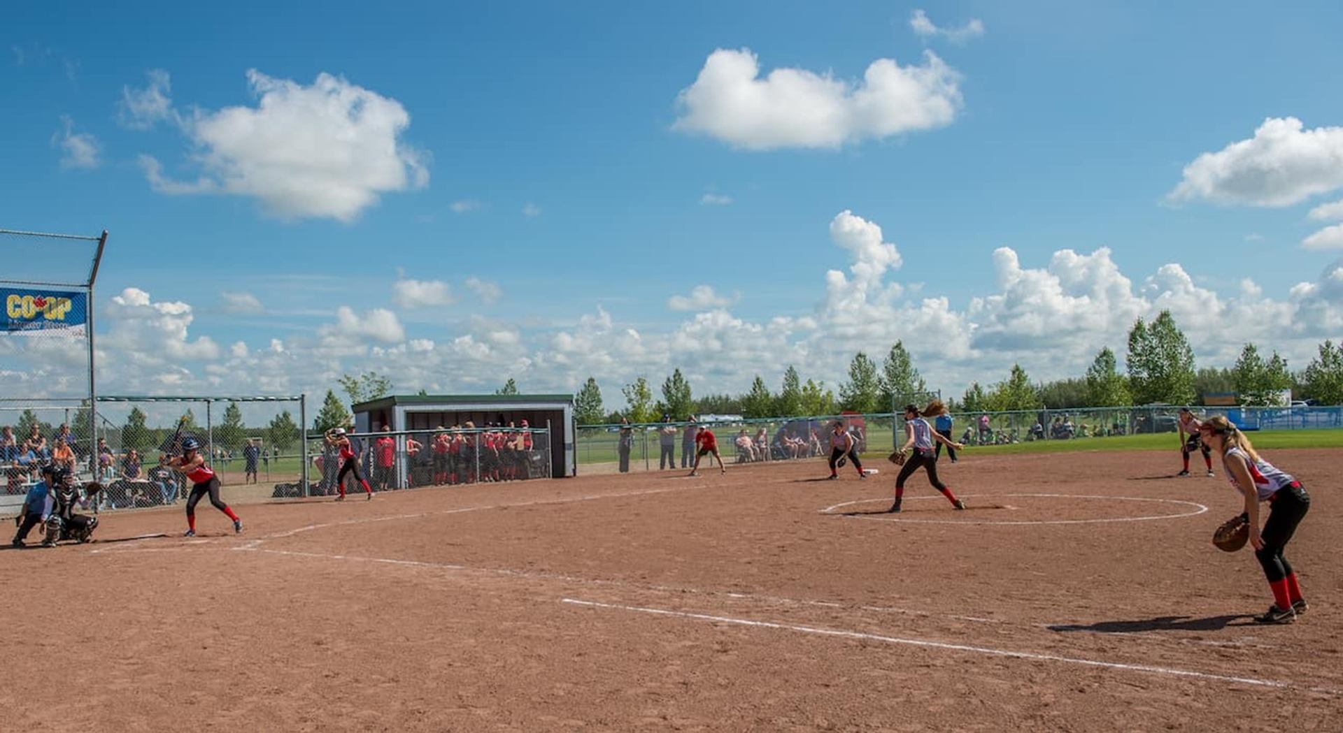 Softball players on a dirt field during a game, with spectators behind fences.