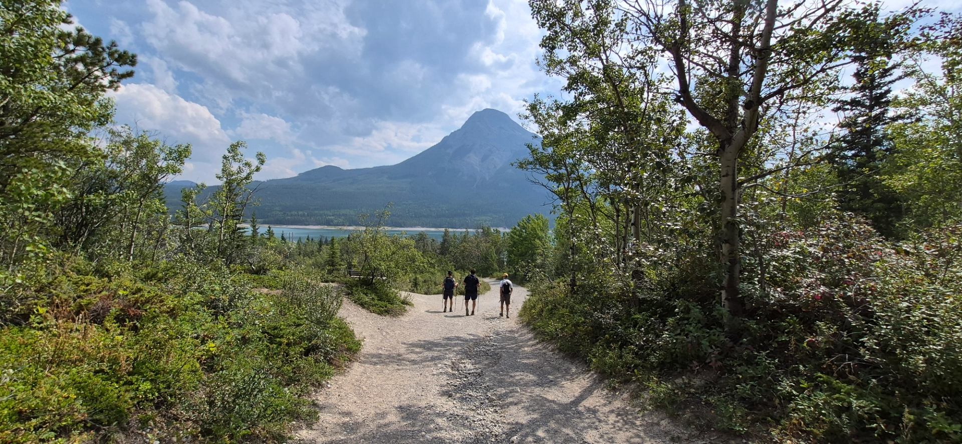 Hikers descend a forest trail toward a blue lake and mountain.
