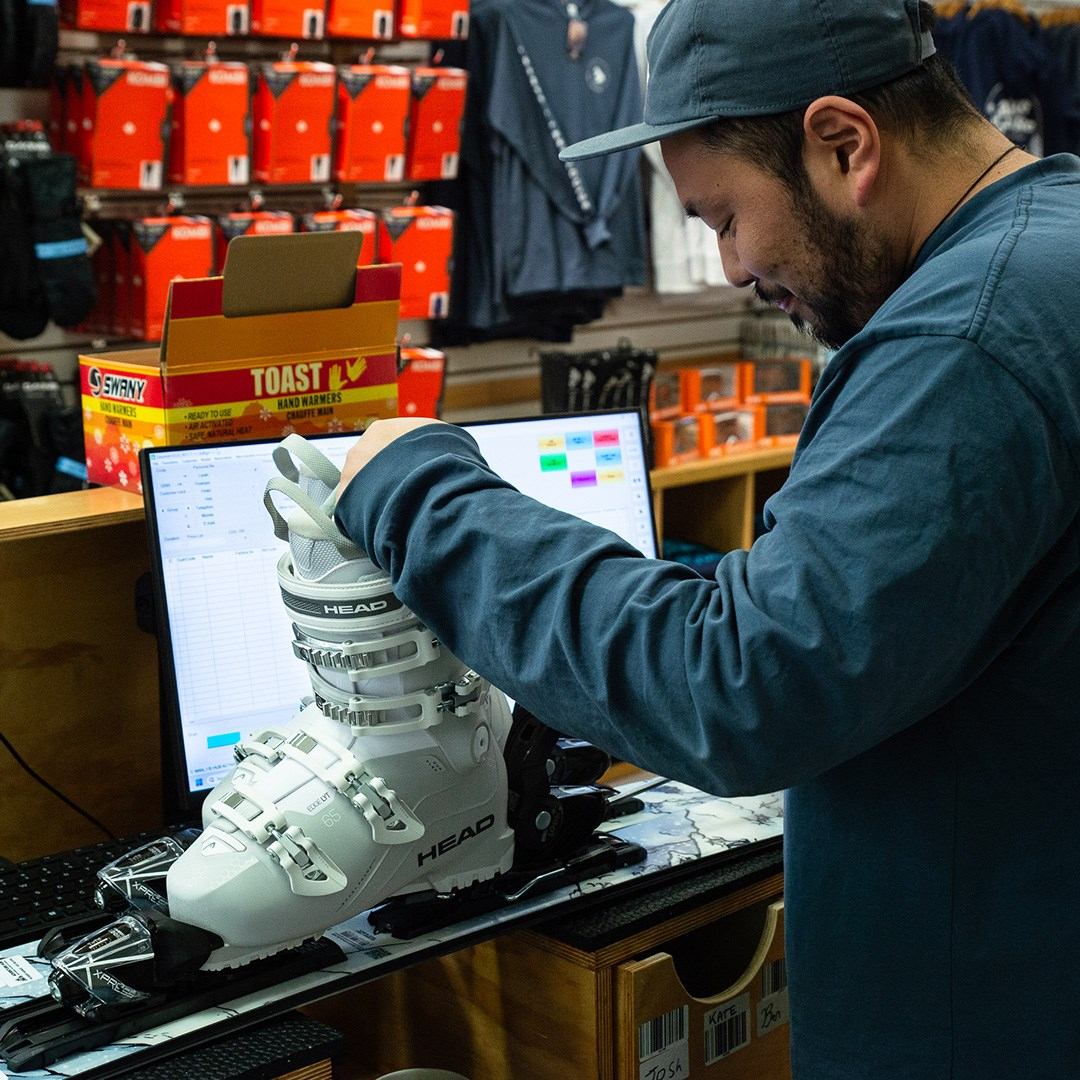 Person fitting a ski boot at a counter with tools, monitor, and gear in a workshop setting.