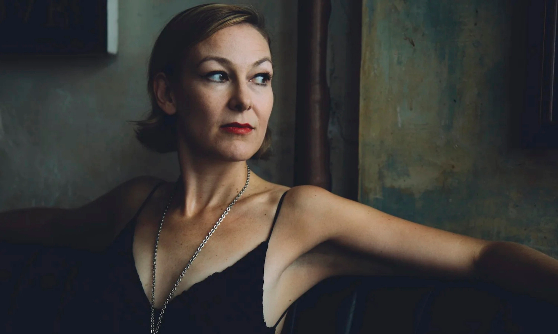 Person in black camisole sits with arms outstretched against a textured wall.