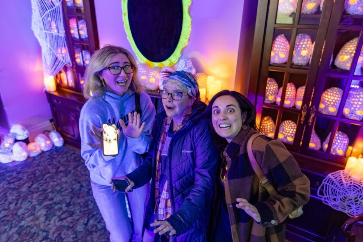 Three people pose among glowing lanterns, purple lighting and decorative spiderwebs.