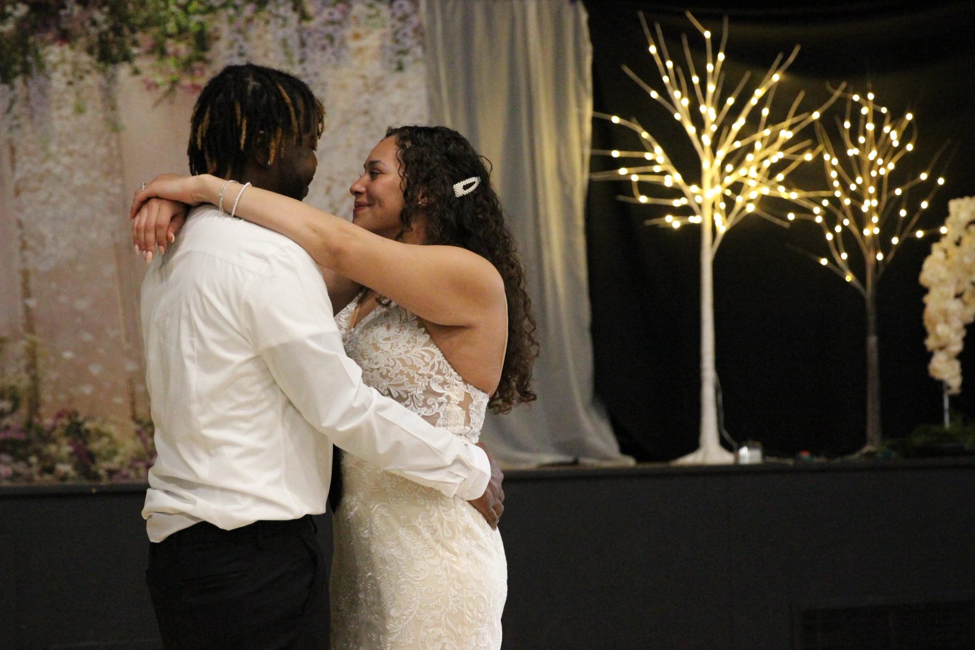 Couple dancing closely in formal attire beside decorative lit trees and draped backdrop.