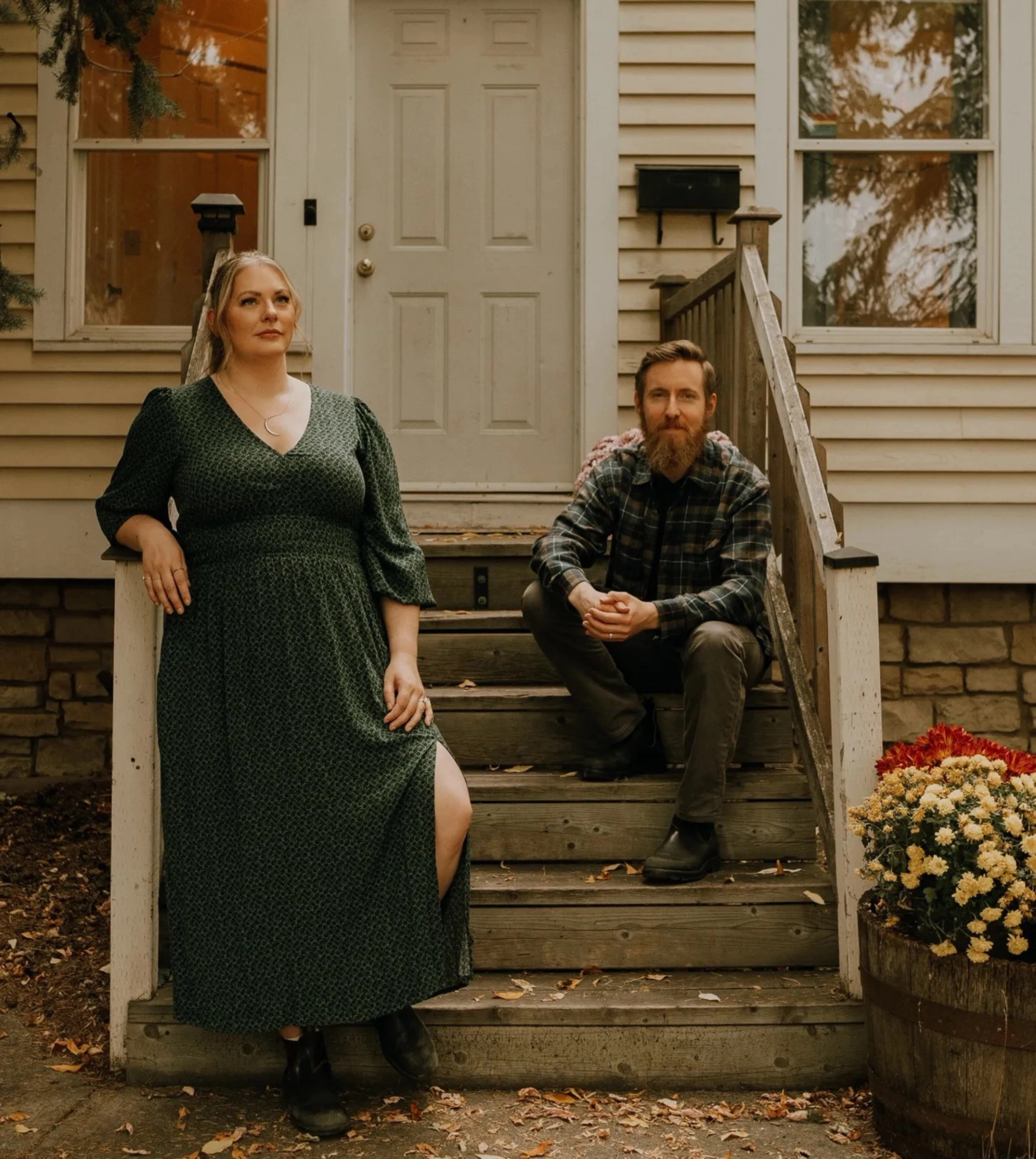 Two adults on wooden house steps, one standing, one sitting, beside potted flowers.