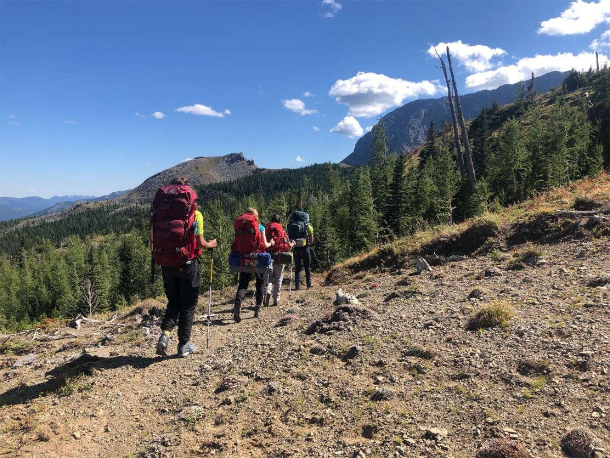 Hikers with large backpacks walk along a rocky mountain trail through evergreen forest.