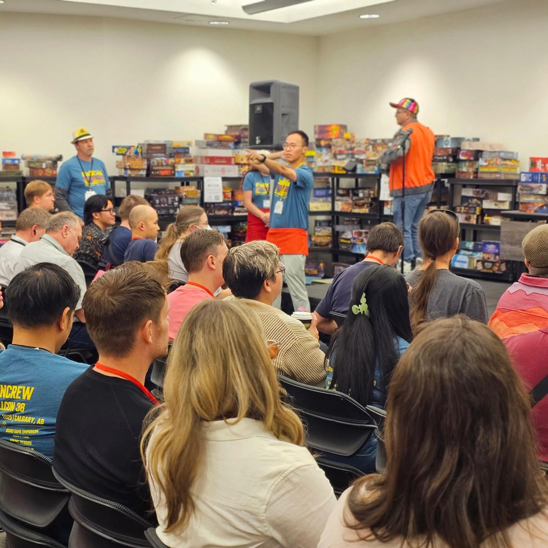 Board game event staff address seated audience in room lined with stacked game boxes.