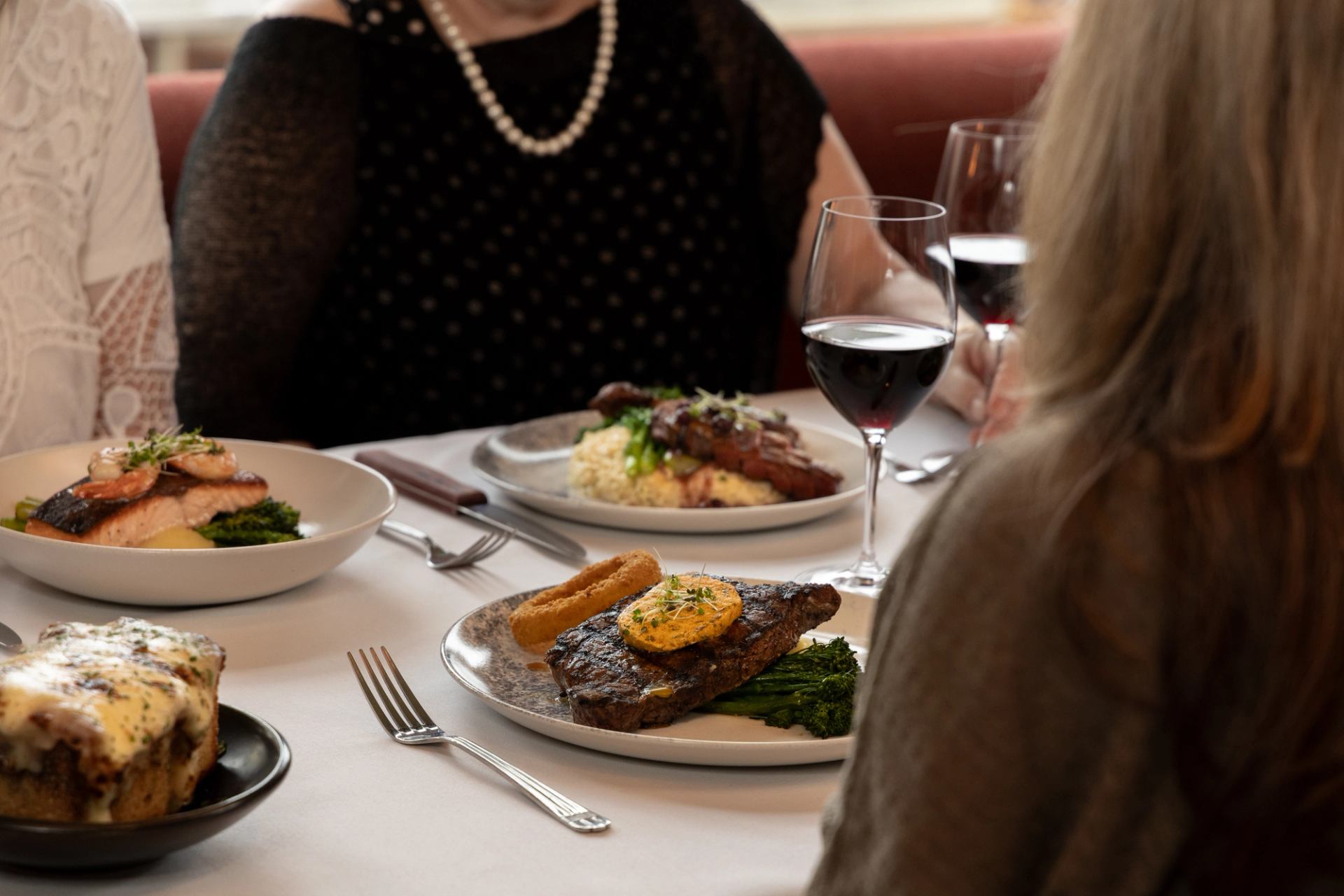 3 people sit around a table with plated seafood, steak, and glasses of wine.