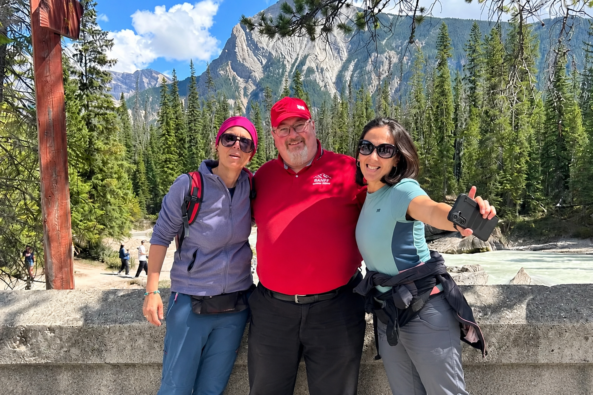Three people pose beside a river with evergreen forest and rocky mountains behind them.