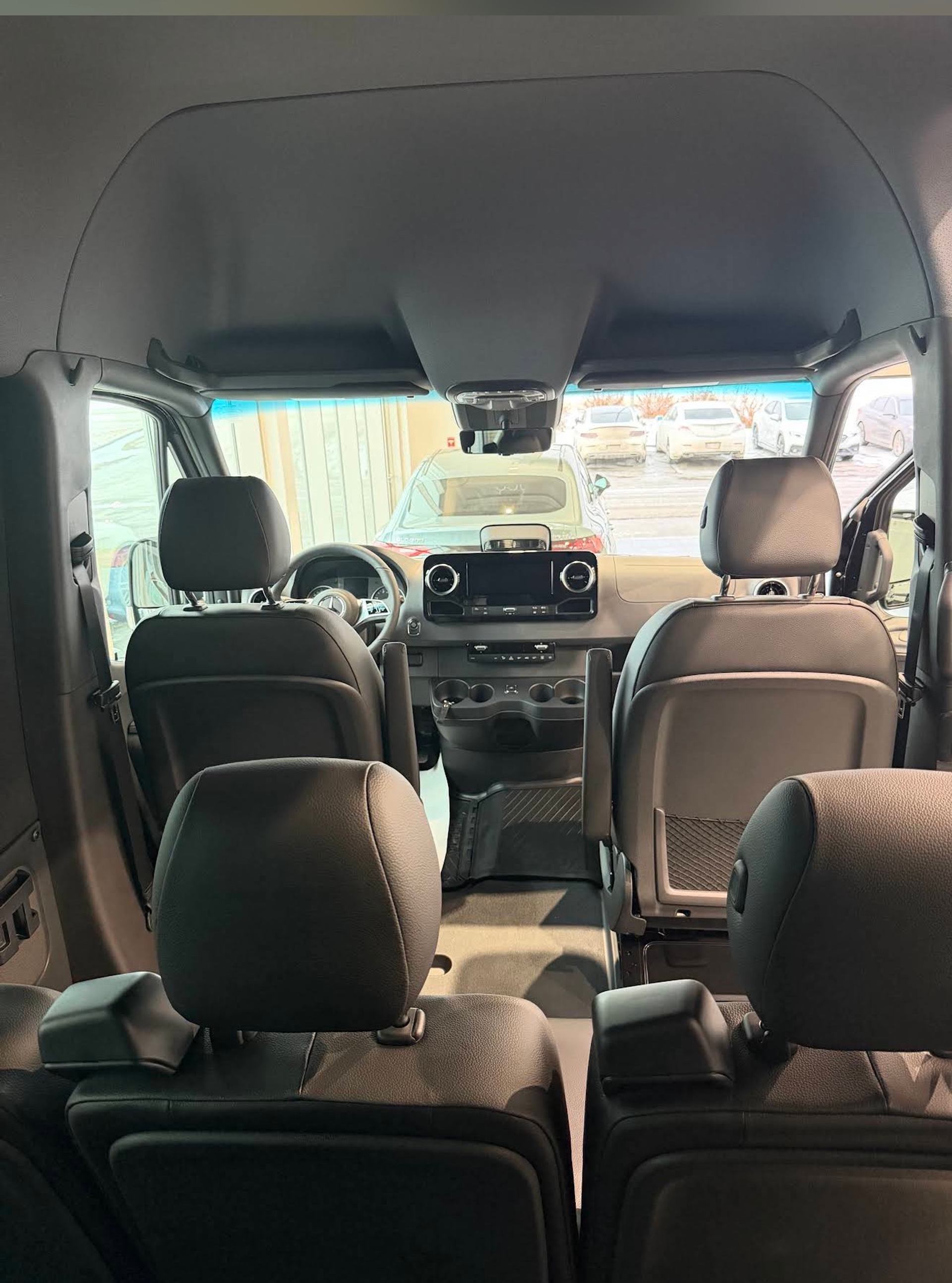 Van interior with multiple rows of seats facing dashboard and parked cars outside.