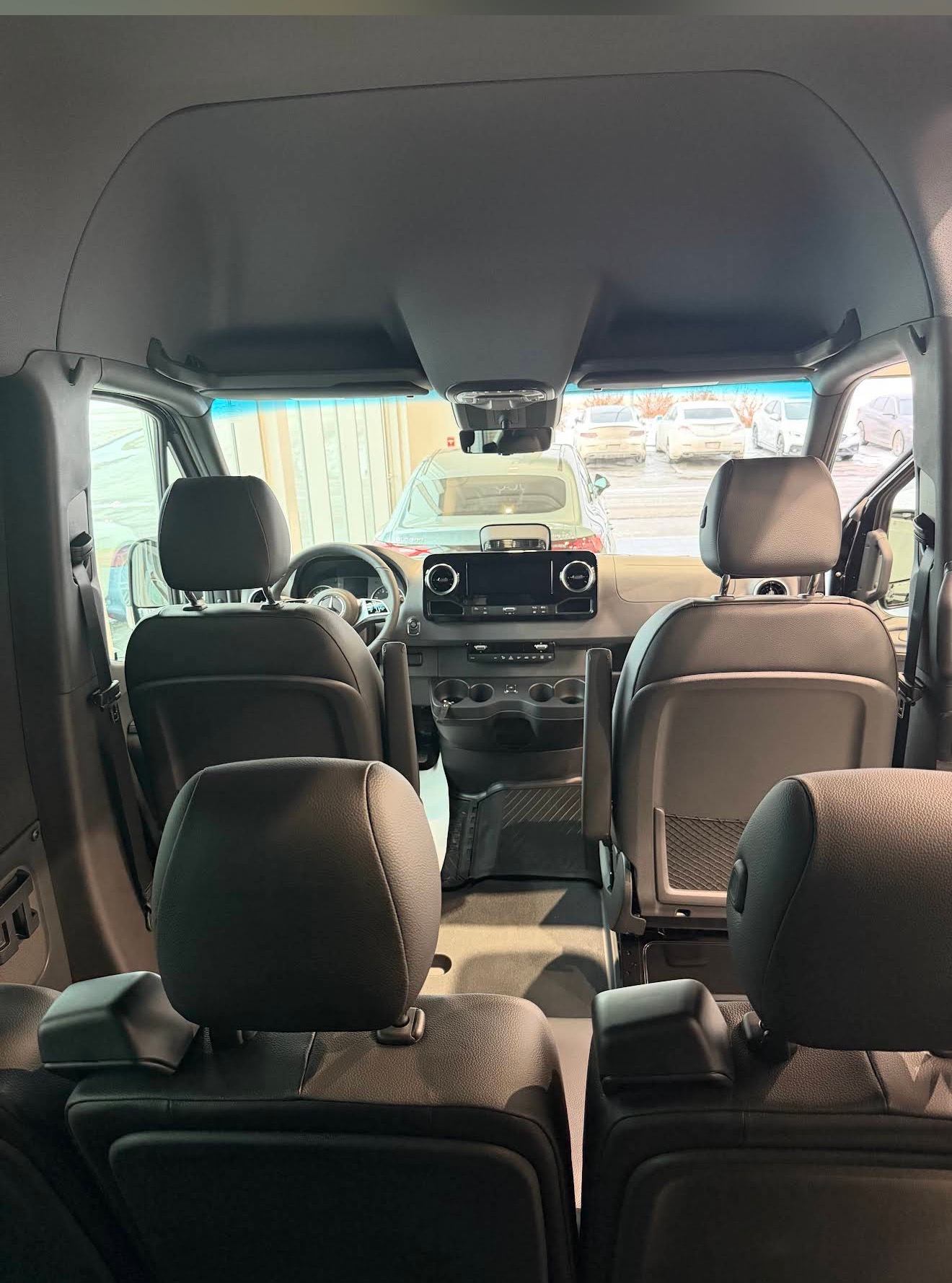 Van interior with multiple rows of seats facing dashboard and parked cars outside.