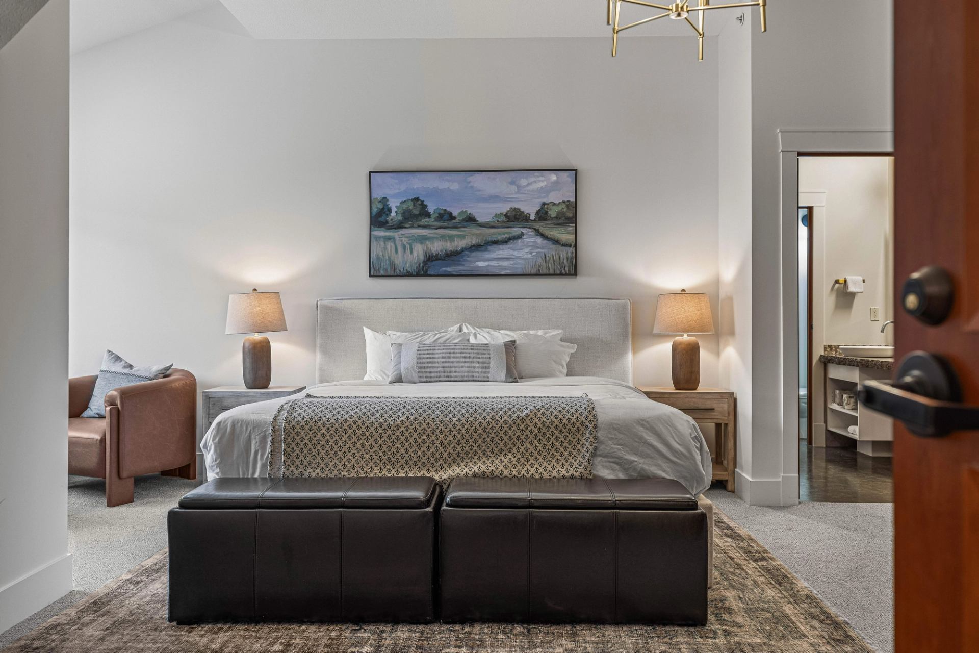 Bedroom with upholstered bed, landscape painting, bedside lamps and leather bench.