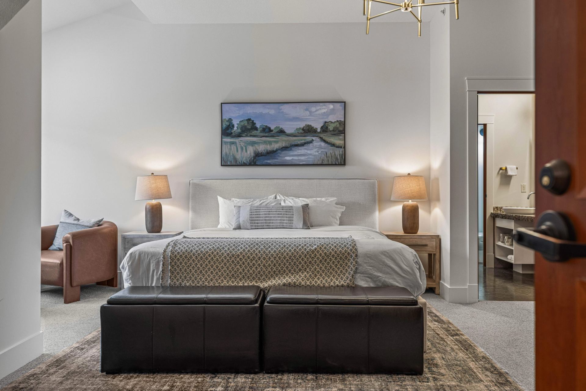 Bedroom with upholstered bed, landscape painting, bedside lamps and leather bench.