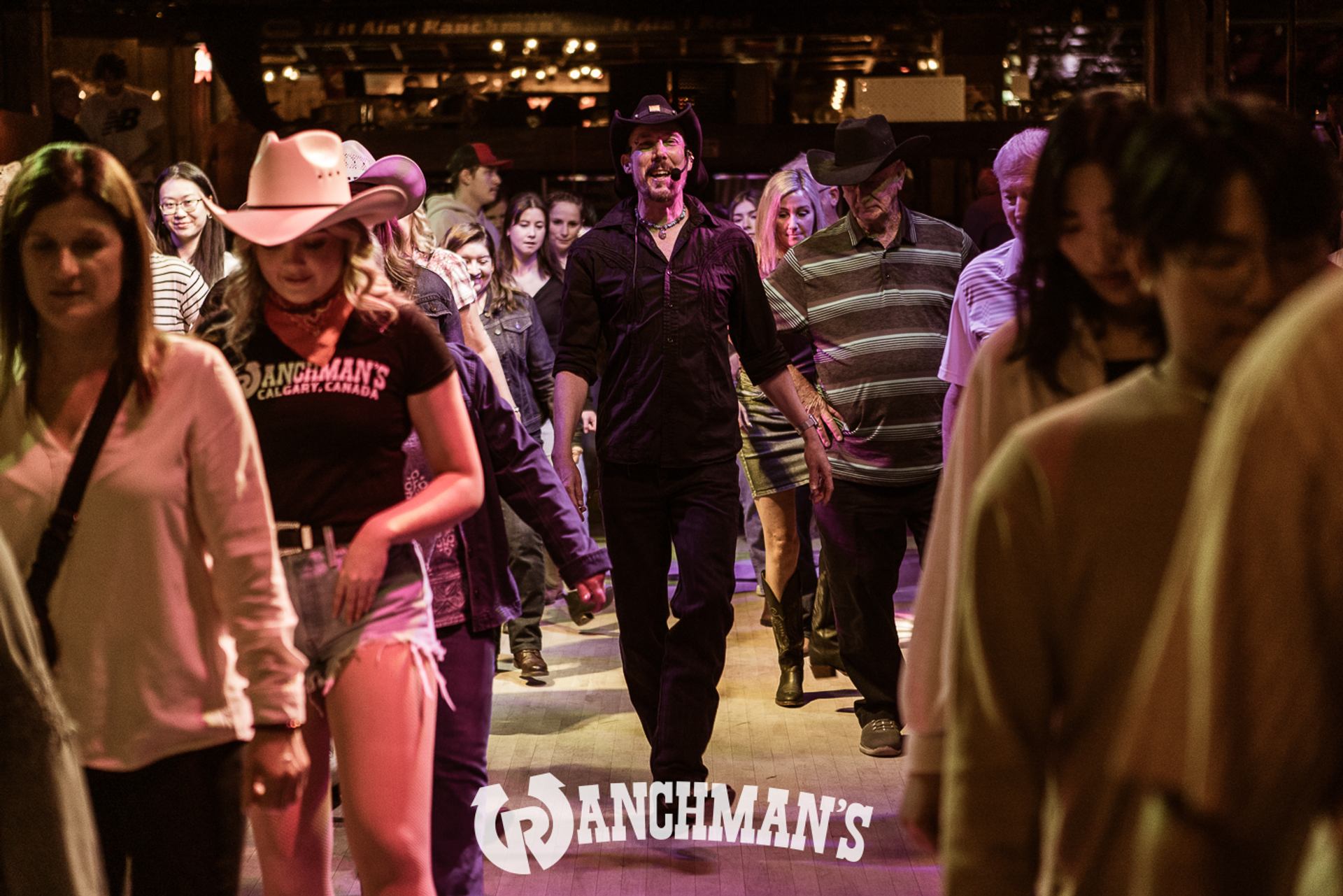 Crowd line dancing indoors at Ranchman’s, with cowboy hats and casual clothing.