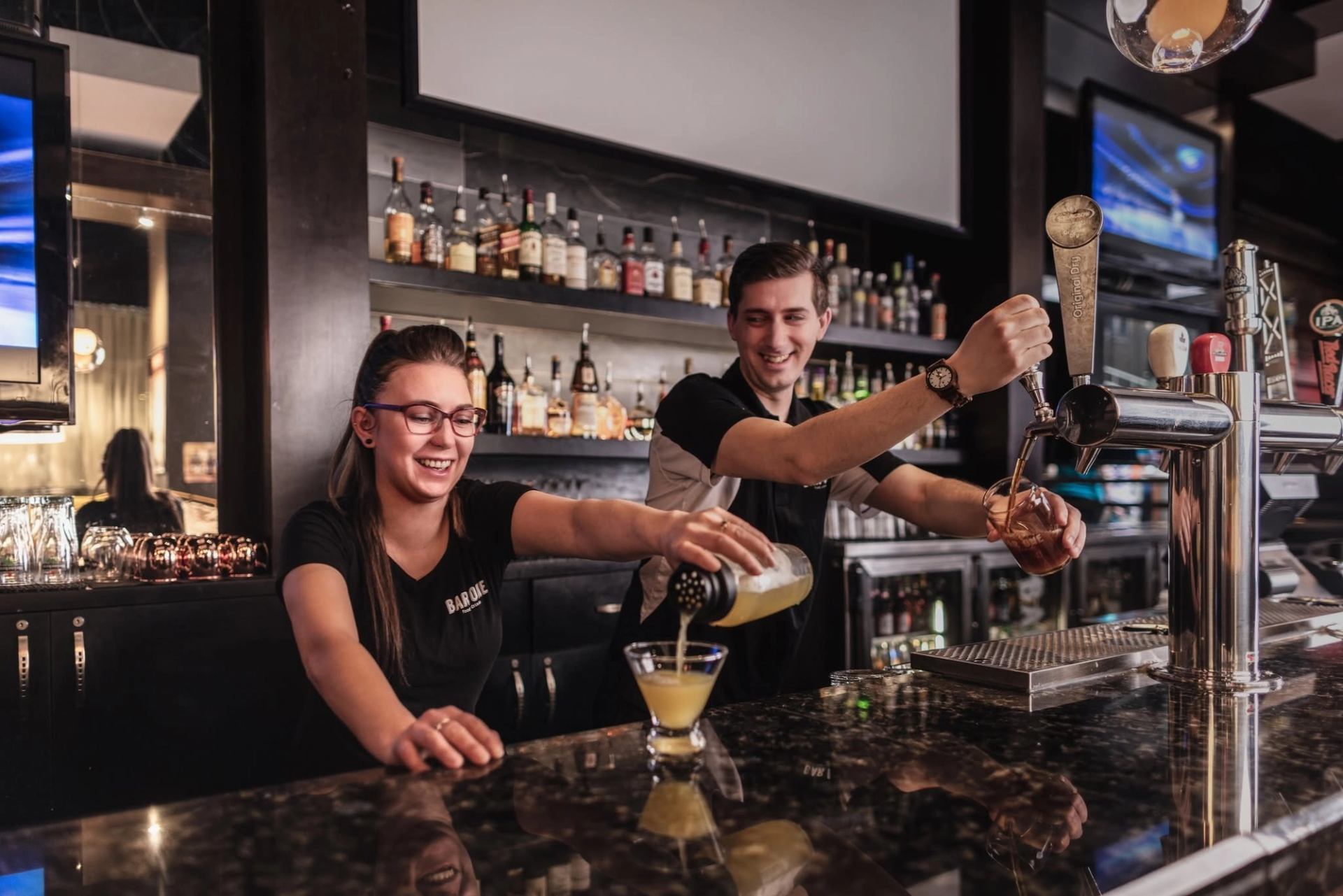 Bartenders pour a cocktail and draught beer behind a bar.