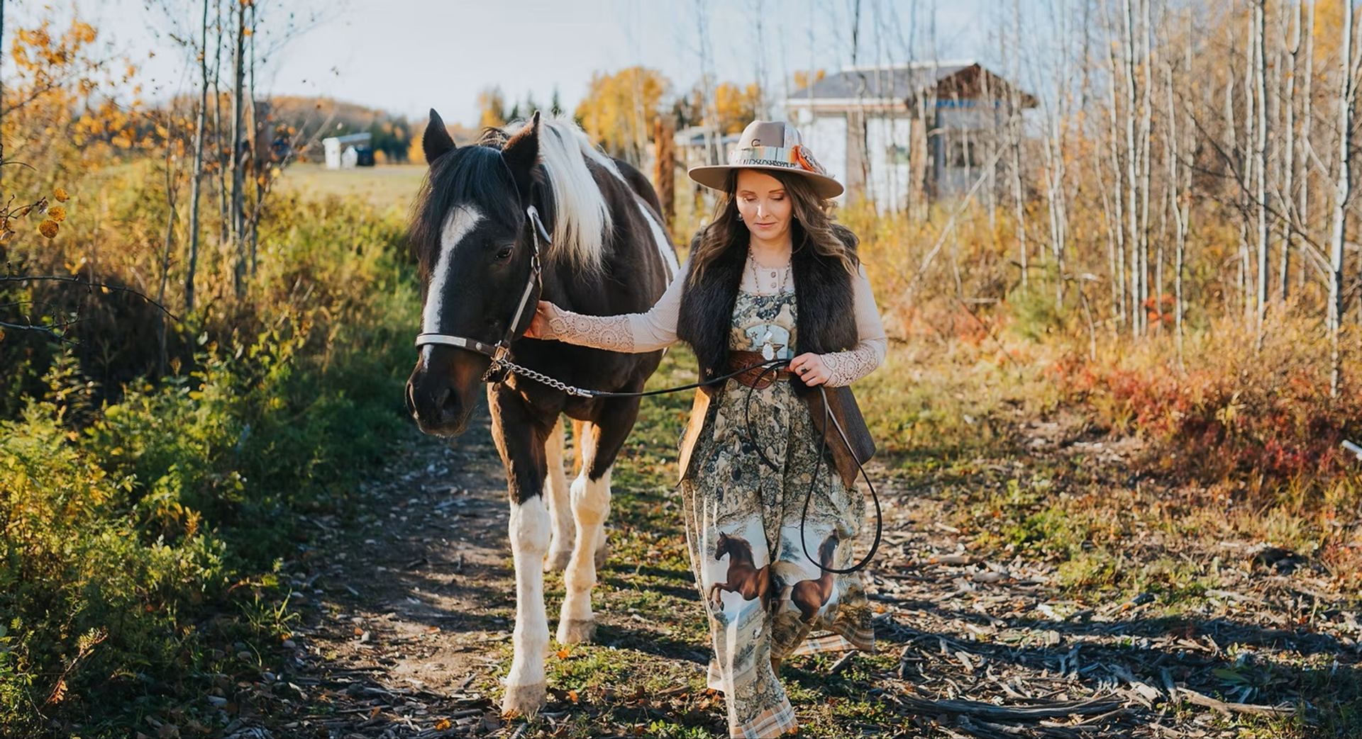 Person in a wide-brimmed hat leads a black-and-white horse along a wooded path.