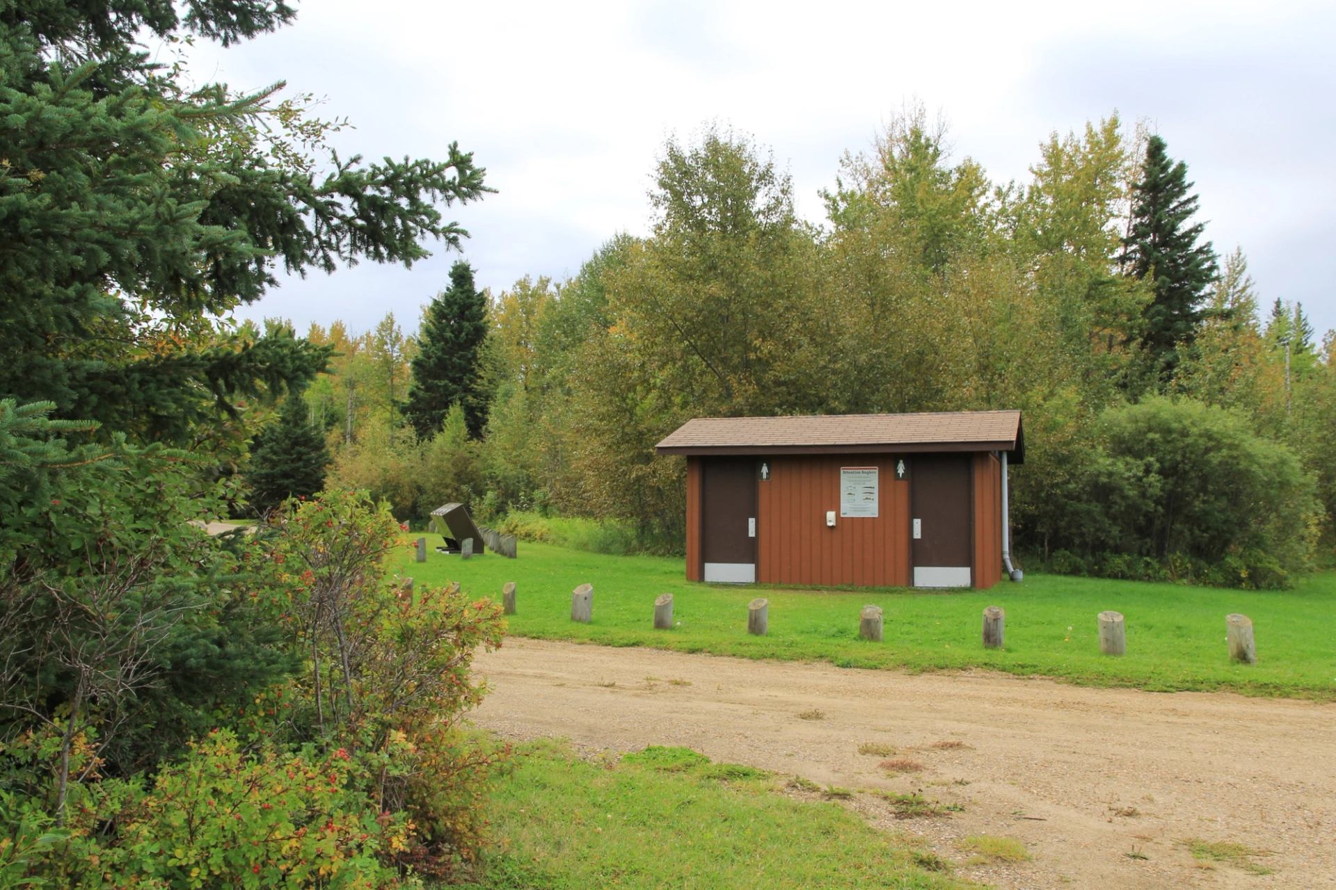 Small brown park building beside gravel road, surrounded by green grass and trees.