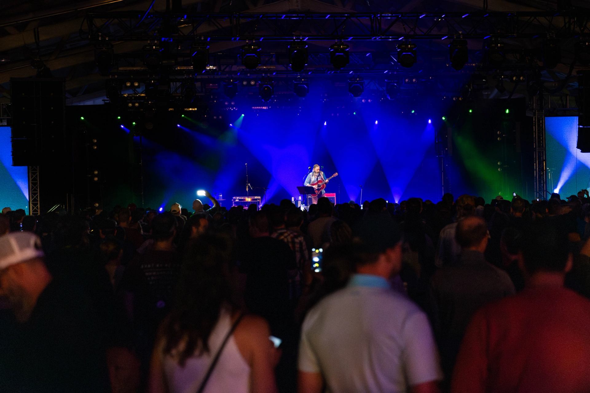 Crowd watches musician playing guitar on stage under blue and green concert lights.