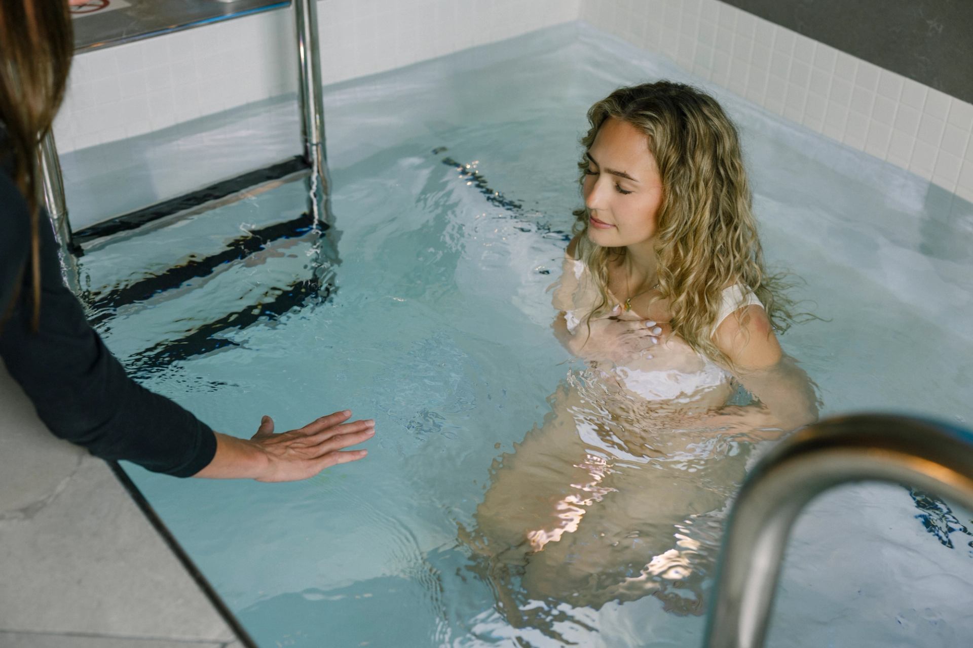 Person in shallow pool holds chest while another person extends hand near ladder.