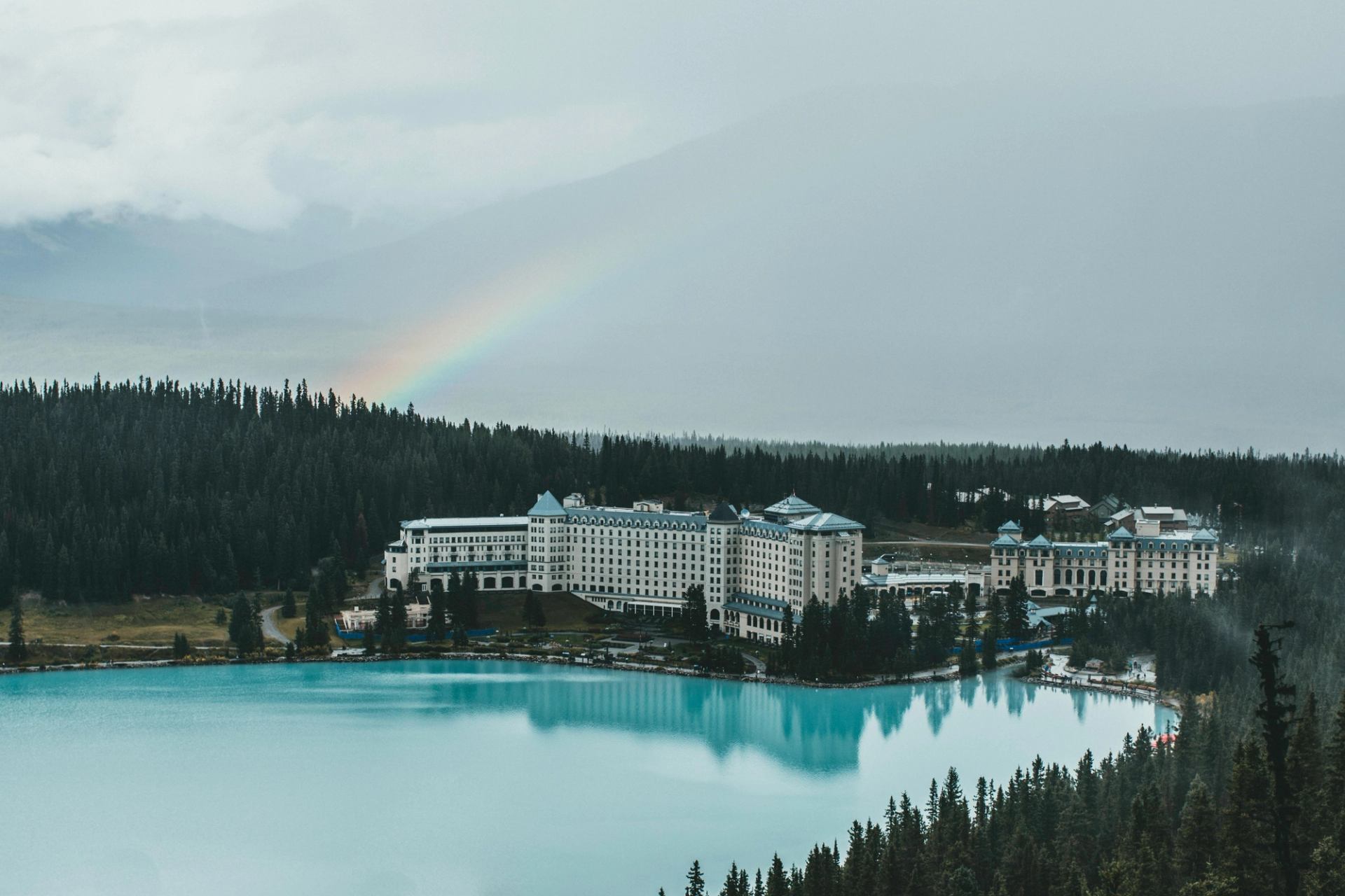 Lakeside hotel with turquoise water and a faint rainbow under cloudy skies.