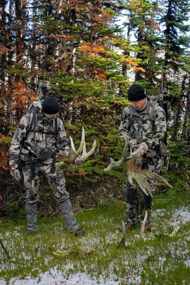 Two people in camouflage hold antlers in a conifer forest with patchy snow.