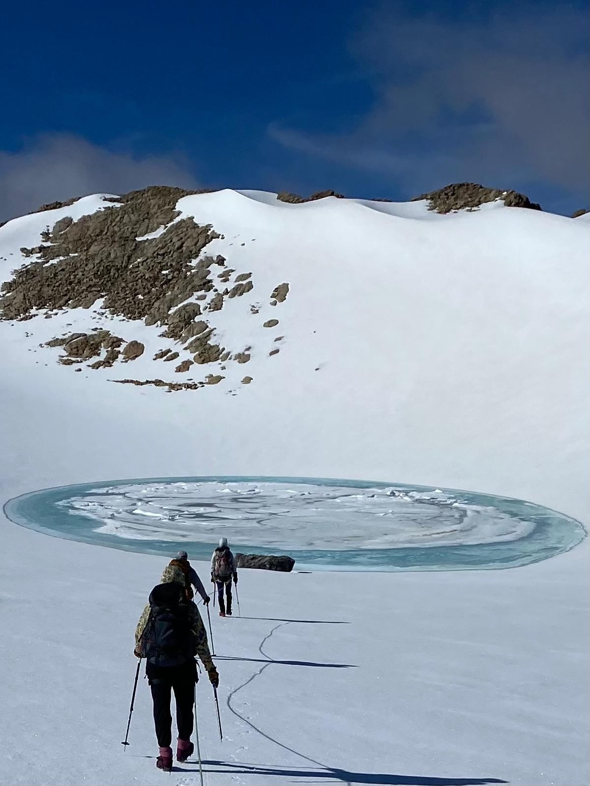 Two hikers approach a circular frozen lake in a snowy mountain landscape.