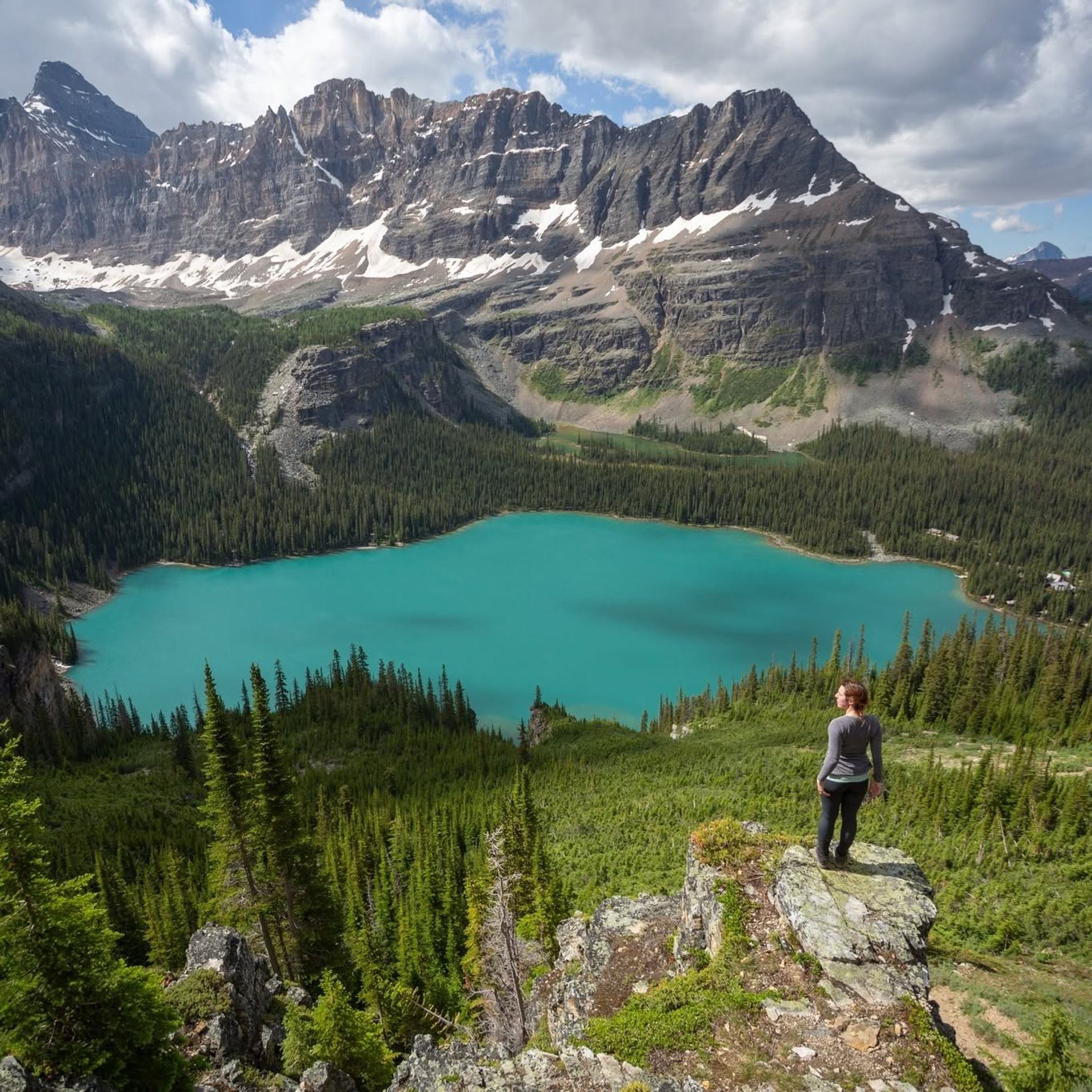 Hiker overlooks a turquoise mountain lake surrounded by evergreen forest and snow-streaked peaks.