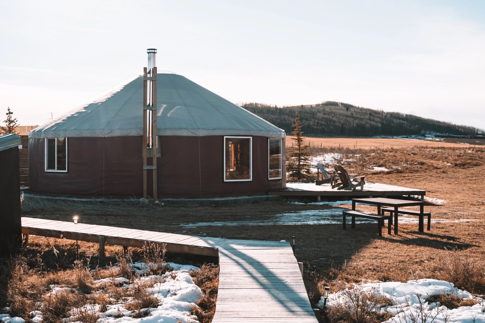 Round cabin with chimney, wooden walkways and chairs amid patchy snow and fields.