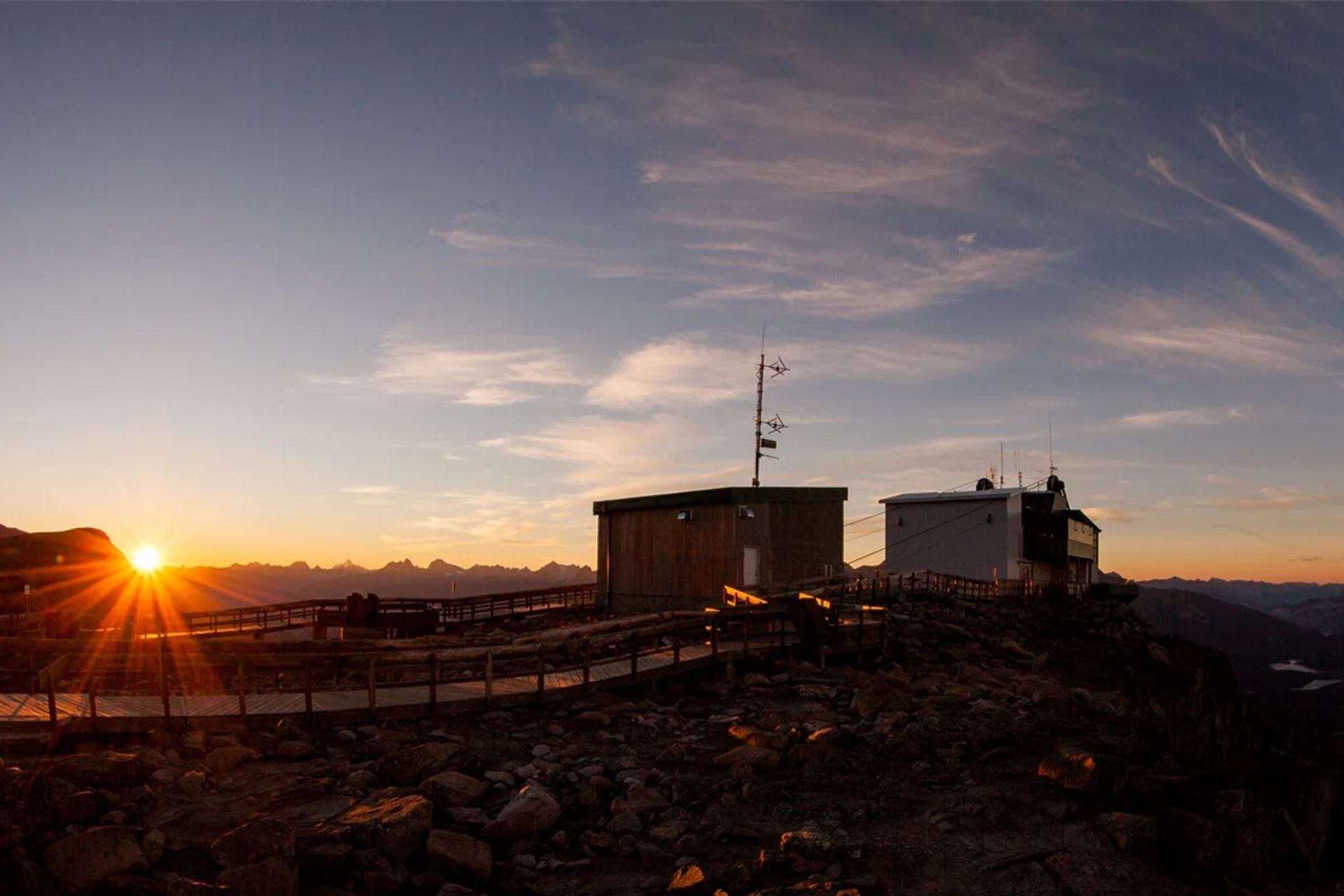 Mountain summit buildings with antennas and boardwalk at sunrise over distant peaks.
