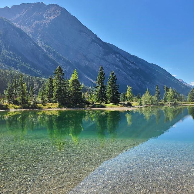 A tranquil lake reflects lush evergreen trees and a towering mountain under a clear blue sky, creating a serene and peaceful natural landscape.