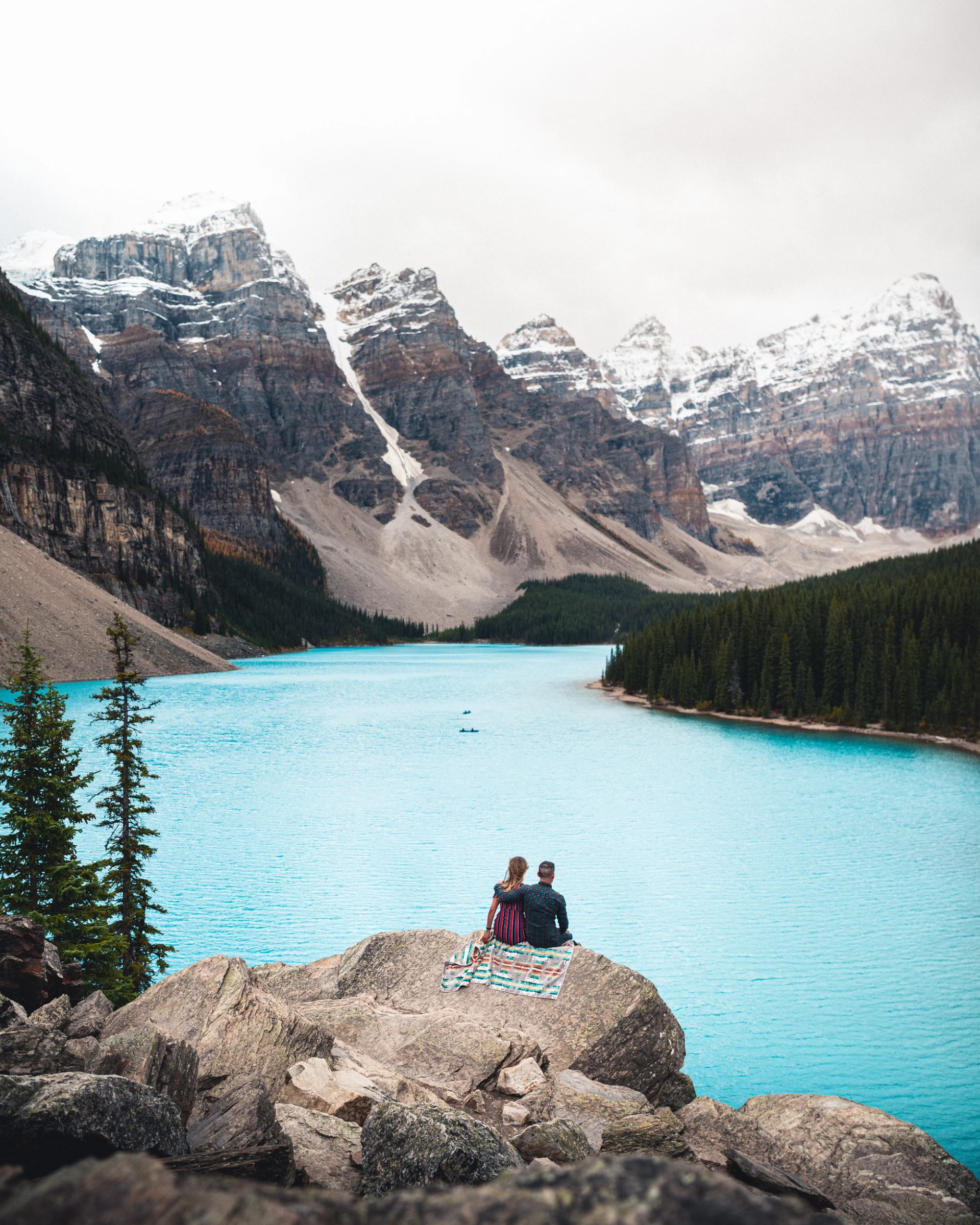 Two people sit on rocks overlooking turquoise lake and snow-capped mountain peaks.