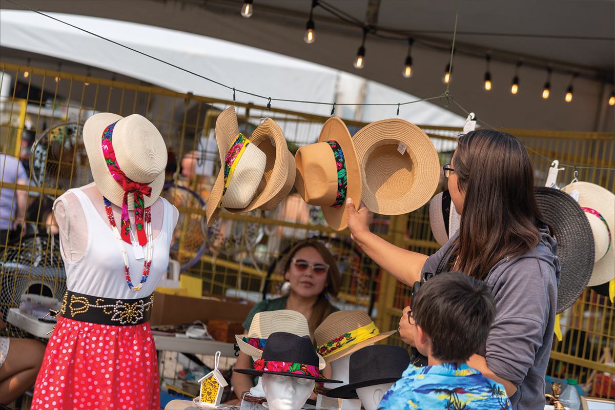 Shoppers browse straw hats with colourful bands at an outdoor market stall display.