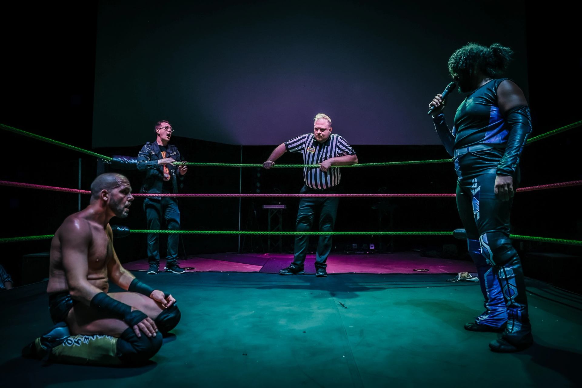 Wrestler with microphone faces seated opponent as referee and announcer watch.