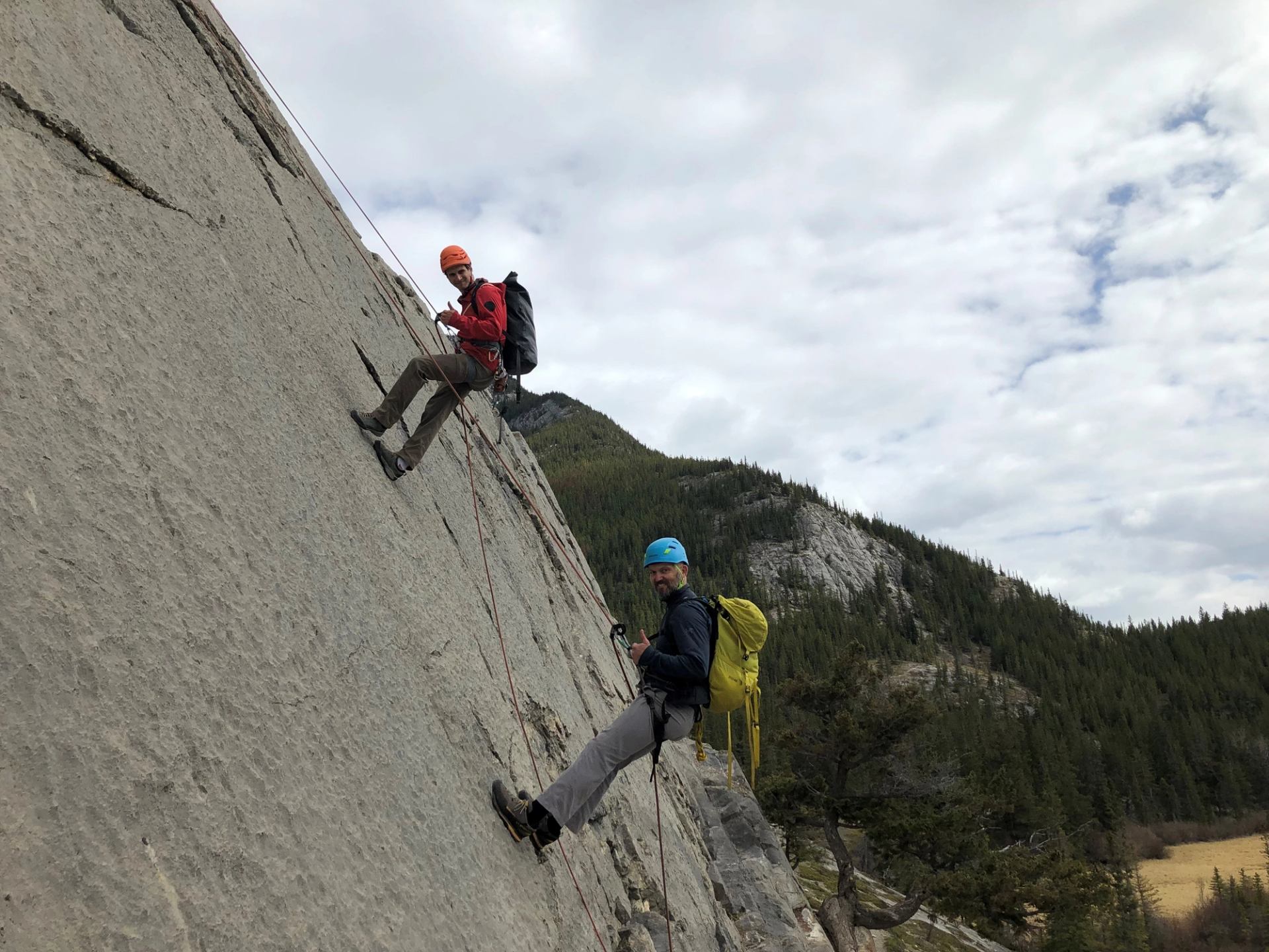 Two climbers with helmets and backpacks ascend steep rock face in cloudy mountainous terrain, evoking adventure and focus.